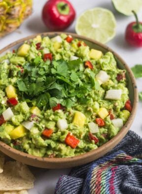 Large bowl filled with pineapple jicama guacamole with a blue towel, chips, limes, and cherry peppers around it