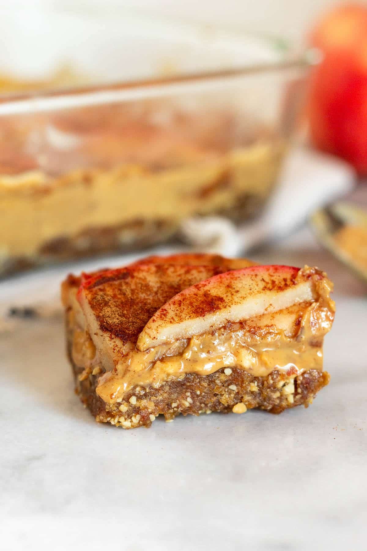 Close up of an apple peanut butter bar sitting on a white counter. Behind it is a pan of more bars, a spoon with peanut butter on it, and an apple.