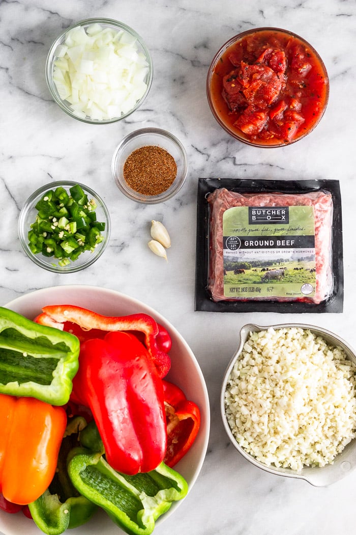 White counter filled with a bowl of halved bell peppers, bowl of chopped jalapeno, bowl of spices, bowl of chopped onion, bowl of canned tomatoes, package ground beef, garlic cloves, and bowl of cauliflower rice.