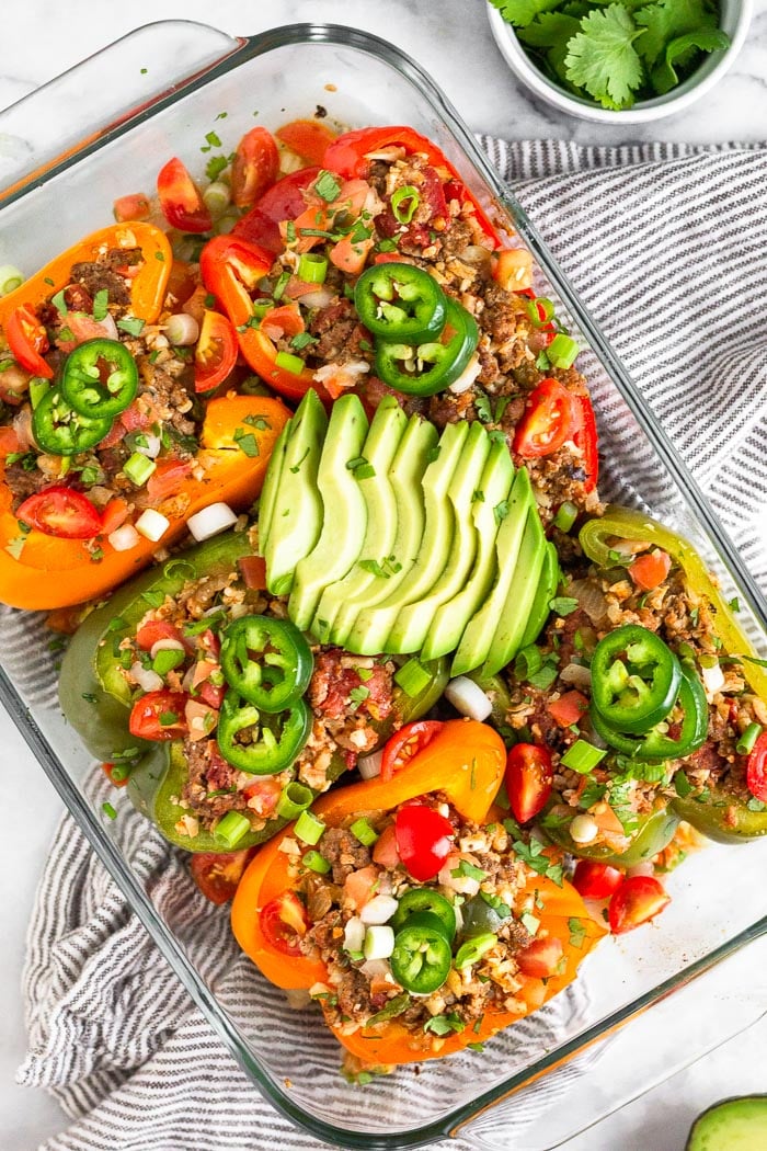 Overhead shot of a glass baking dish filled with stuffed peppers with ground beef and rice. They are topped with sliced avocado, jalapeno, cilantro, and green onion. The pan is sitting on a striped towel with a small dish of cilantro next to it.