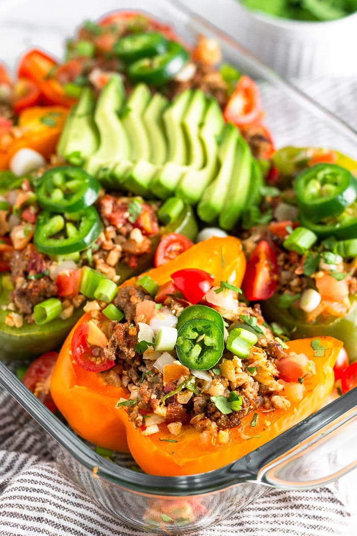 Baking dish filled with Whole30 stuffed peppers with beef and cauliflower rice. They are topped with avocado, jalapeno, cilantro, diced tomatoes, and green onion. The dish is sitting on a striped towel.