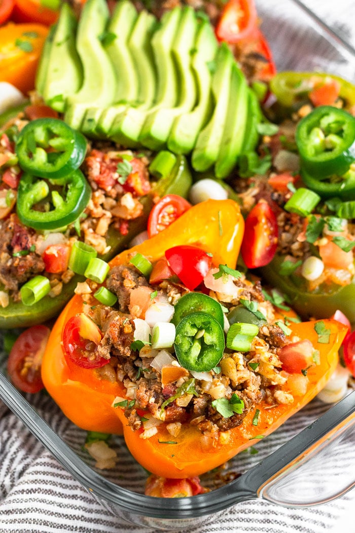 Glass dish filled with beef and rice stuffed peppers topped with sliced jalapeno, green onion, diced tomatoes, cilantro, and sliced avocado.