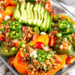 Baking dish filled with Whole30 stuffed peppers with beef and cauliflower rice. They are topped with avocado, jalapeno, cilantro, and green onion. Behind the dish is a small dish of cilantro.