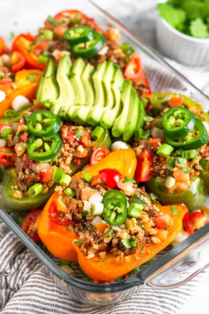 Baking dish filled with Whole30 stuffed peppers with beef and cauliflower rice. They are topped with avocado, jalapeno, cilantro, and green onion. Behind the dish is a small dish of cilantro.