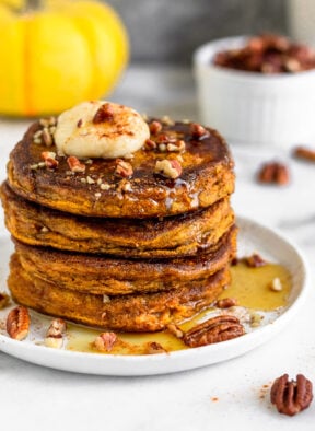 Stack of Protein Powder Pumpkin Pancakes on a white plate with maple syrup, coconut butter, and chopped nuts on top of them. Behind it is a mug, dish of nuts, and a yellow pumpkin.