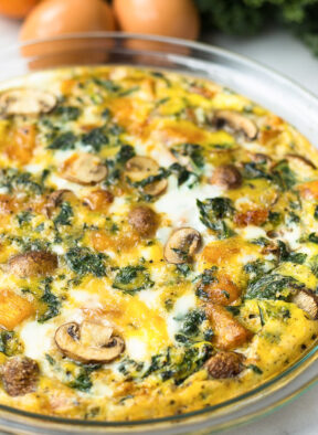 Crustless pumpkin quiche in a pie pan with eggs, pumpkin, and kale behind it.