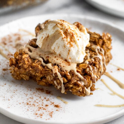 Gingerbread baked oatmeal bar on a plate topped with yogurt, nut butter, and cinnamon. Behind it is another plate and a pan of more baked oatmeal