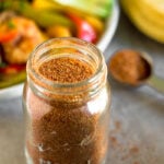 Healthy homemade taco seasoning in a glass jar with a teaspoon behind it filled with seasoning and a plate of fajitas.