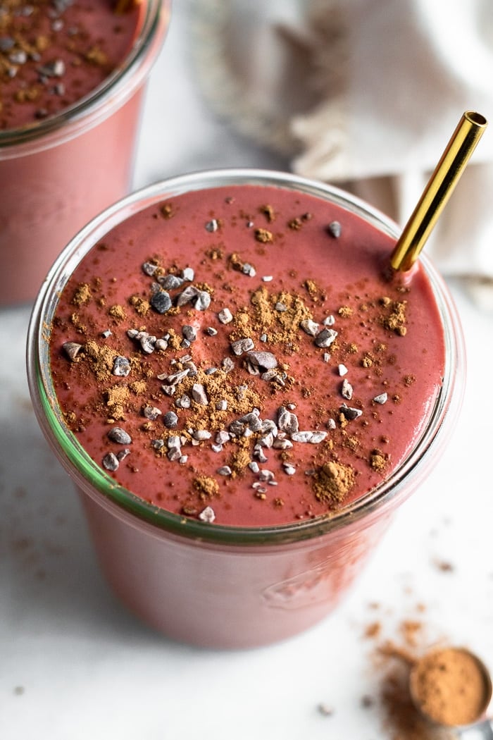 Overshead shot of a beet smoothie topped with cacao nibs and cacao powder. Também contém uma palha de ouro. Ao lado está outro batido e uma colher de chá cheia de cacau em pó.