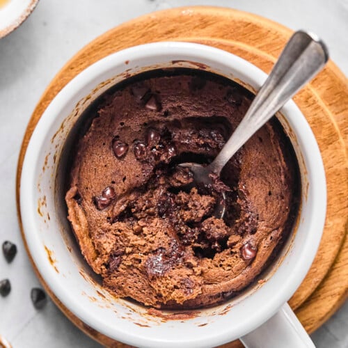 Overhead shot of a protein mug cake with a spoon in it with some of the middle scooped out. The mug is sitting on a small wooden coaster and around it are some mini chocolate chips.