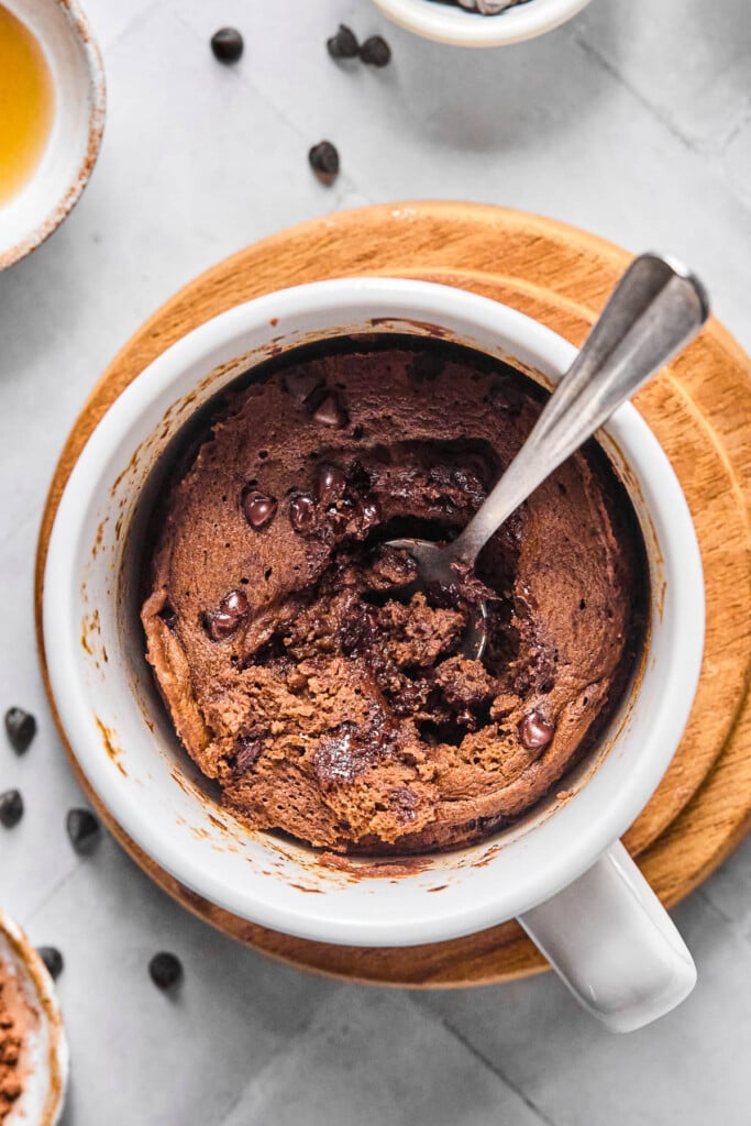 Overhead shot of a protein mug cake with a spoon in it with some of the middle scooped out. The mug is sitting on a small wooden coaster and around it are some mini chocolate chips.