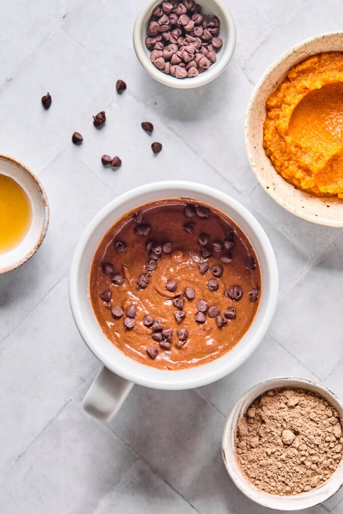Overhead shot of a white mug with a chocolate batter with mini chocolate chips in it. Around the mug is a bowl of pumpkin puree, a bowl of chocolate protein powder, a bowl of maple syrup, and a bowl of mini chocolate chips with some around the bowl.