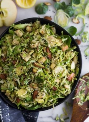 Big bowl with apple & shaved brussels sprouts salad surrounded by half an apple, half a lemon, brussels sprouts leaves, and almonds