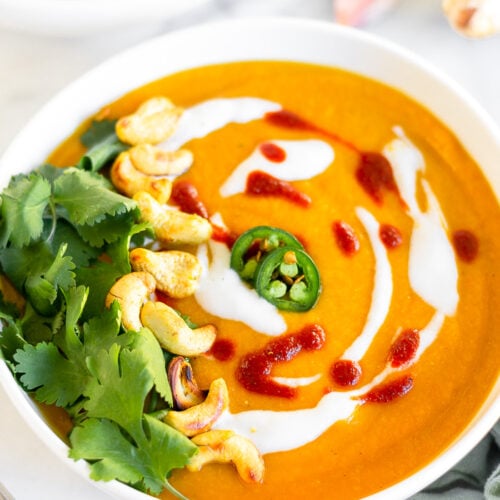 Bowl of curried butternut squash soup topped with coconut milk, hot sauce, sliced jalapenos, cashews, and cilantro. Behind it is another bowl and a clove of garlic and fresh ginger.