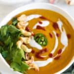 White bowl on a marble counter with curry cauliflower butternut squash soup garnished with cilantro, cashews, coconut yogurt, hot sauce, and jalapeños