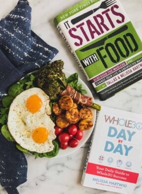 Plate with greens, fried eggs, broccolini, sautéed plantains, and cherry tomatoes next to two Whole30 books.