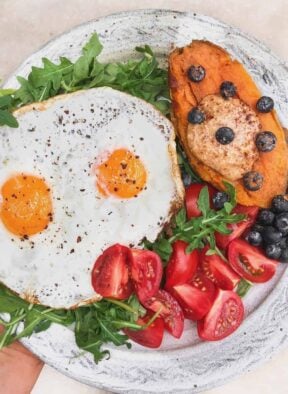 Plate with two fried eggs, half a sweet potato with almond butter and blueberries on it, and tomatoes all over greens