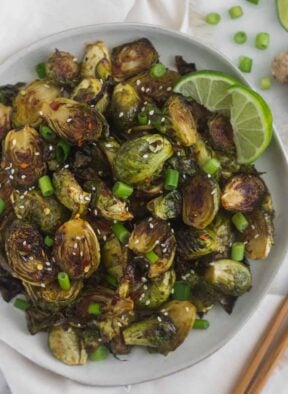 Plate of miso glazed brussels sprouts garnished with sesame seeds, green onion, and lime wedges surrounded by chopsticks and more green onions and limes