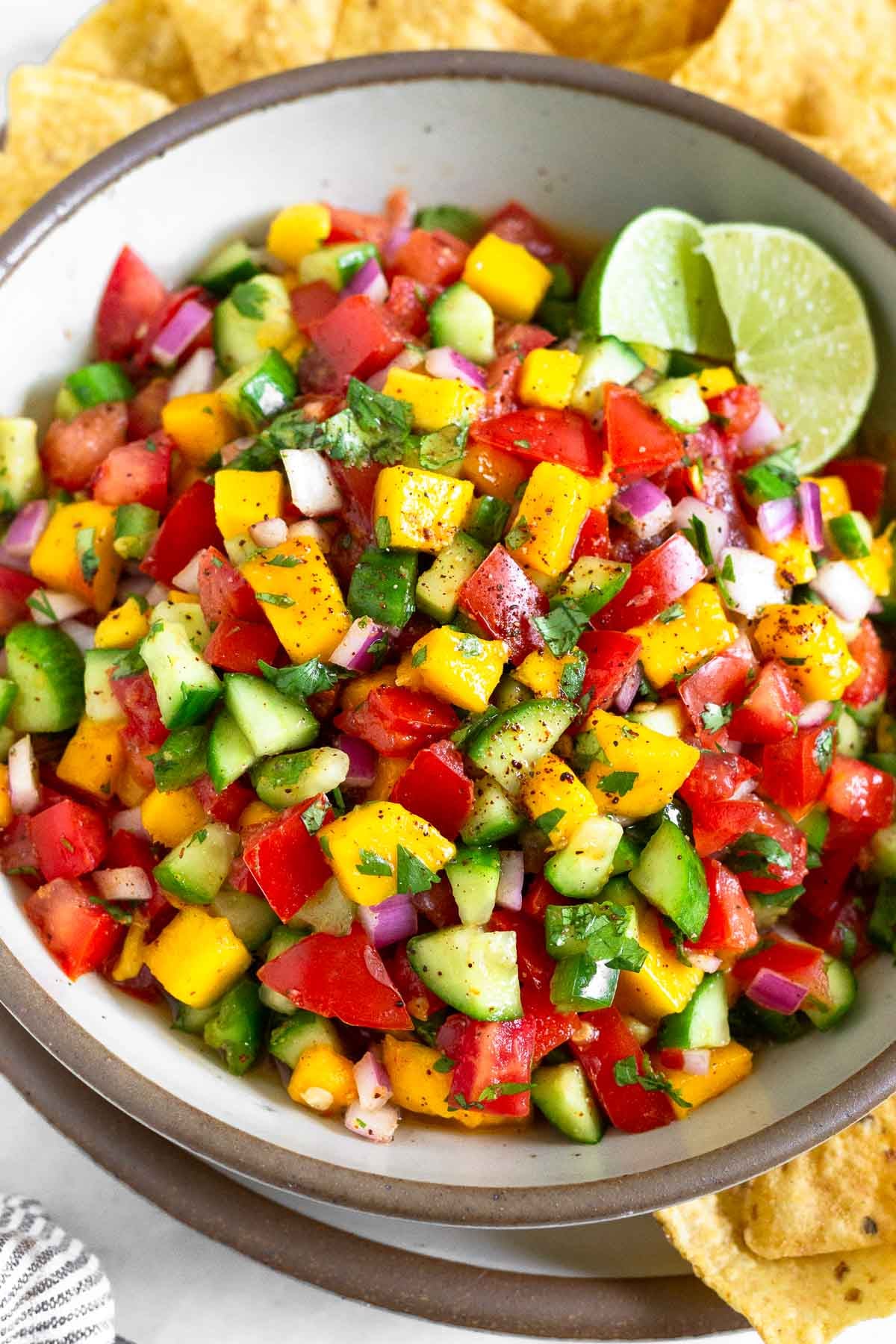 Mango cucumber salsa in a white bowl garnished with two lime wedges. The bowl is sitting on a large plate with tortilla chips on it.