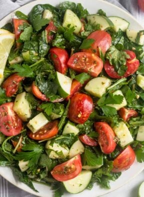 Large white plate filled with Chopped Tomato Cucumber Kale Salad surround by salad tongs, sliced cucumber, and tomatoes