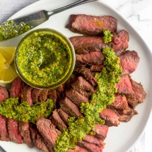 White plate with cut up grilled steak with pistachio pesto on top of it along with a jar of pistachio pesto and sliced lemon