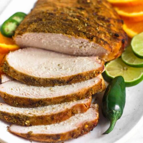 Citrus grilled turkey breast with a few pieces cut laying on a plate next to some sliced limes, oranges, and a jalapeño.