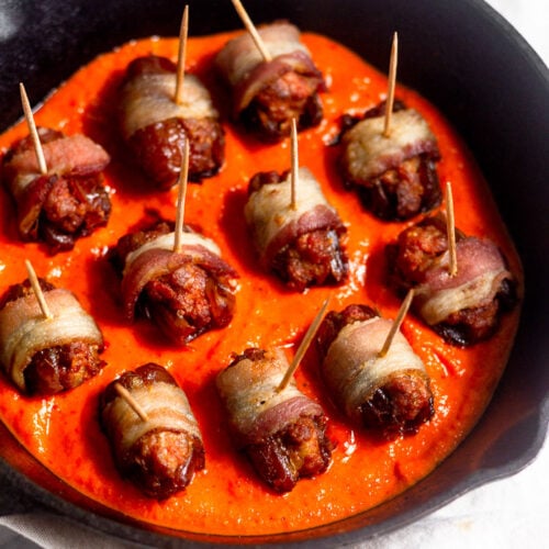 Small cast iron skillet bacon-wrapped chorizo-stuffed dates sitting on top of red pepper sauce.