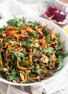 Large white bowl filled with a paleo kale salad with butternut squash and pomegranates. It also has walnuts and pumpkin seeds in it. Behind the bowl is a quarter of a pomegranate, half a lemon, and a small bowl of dressing further behind them.