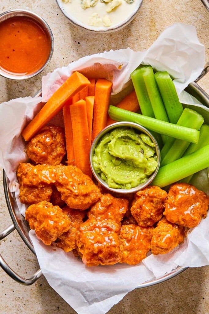 Overhead shot of a large dish of boneless buffalo wings with carrot sticks, celery sticks, and a small ramekin of guacamole for dipping. Next to the dish is a small bowl of buffalo sauce and a small bowl of blue cheese dressing.