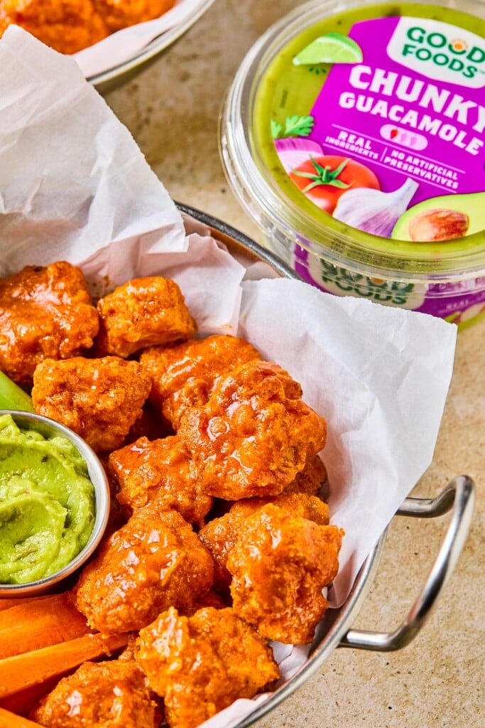 Dish of air fryer boneless buffalo wings with a small ramekin of guacamole and carrots sticks in it. Behind the dish is a container of Good Foods Chunky Guacamole and another dish of wings.