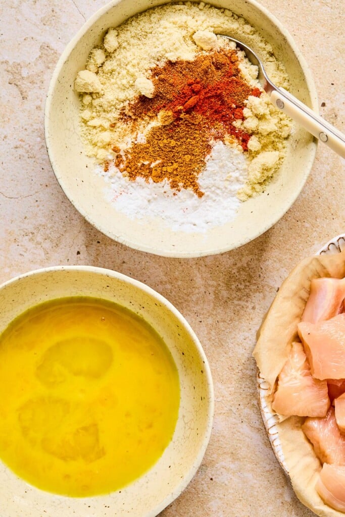 Overheat shot of a bowl of two kinds of flours and spices with a whisk in it before it is mixed together. Next to it is a plate of raw cubed chicken and a bowl with bowl with whisked eggs in it.