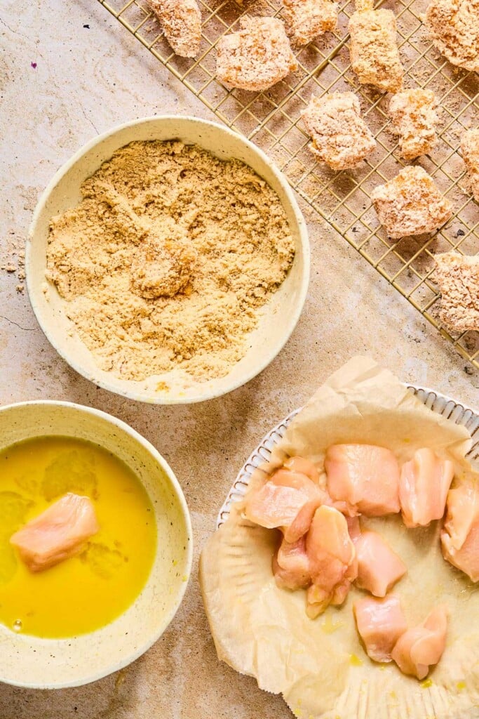 Assembly line of breading chicken with a plate of cubed raw chicken bites, a bowl of whisked eggs with a piece of chicken in it, a bowl of a flour mixture, and a wire rack with pieces of breaded chicken on it.