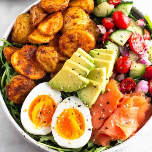 Breakfast bowl filled with greens, roasted potatoes, tomato and cucumber salad, smoked salmon, sliced avocado, and a hard-boiled egg and topped with everything but the bagel seasoning. Next to it is half an avocado.