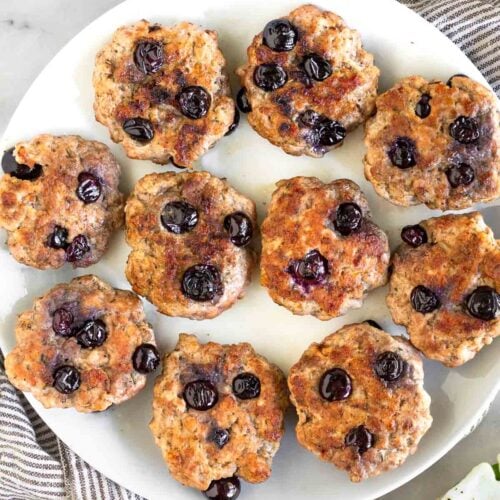 A white plate of blueberry breakfast sausage patties. They plate is sitting on a kitchen towel and next to it is a bowl of blueberries.