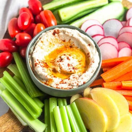 A circular wooden cutting board filled with a bowl of roasted cauliflower hummus surrounded by a bunch of fresh cut fruits and veggies.