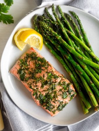 Dill coated salmon and asparagus on a white plate with two lemon wedges next too it. It is on a grey towel with fresh parsley next to it.