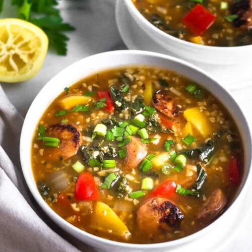 Two bowls of low carb sausage, pepper, and rice soup topped with green onions. A bunch of parsley and half a lemon is behind them and a tan linen is next to the front bowl.