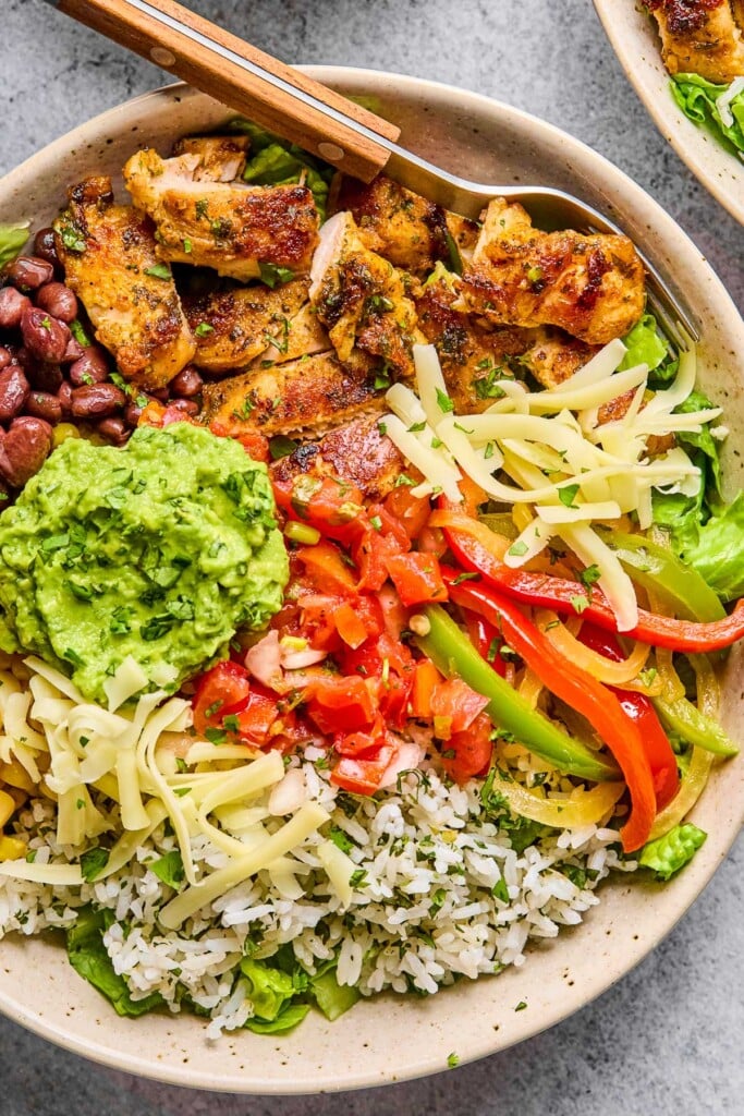 Overhead shot of a white bowl with seasoned sliced chicken, cilantro lime rice, saut&eacute;ed onions and peppers, black beans, lettuce, shredded cheese, salsa, and guacamole. In the bowl is a fork.
