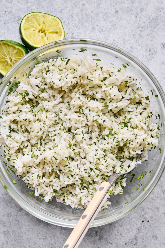 A glass bowl filled with cilantro lime rice with a fork in it. Next to the bowl are two halves of limes that have been juiced.