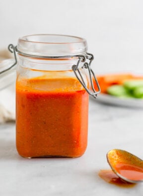 Small jar of homemade Whole30 buffalo sauce recipe. In front of it is a spoon with sauce on it and behind it is a plate of carrots and celery.