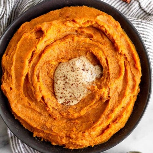Bowl filled with whipped sweet potatoes topped with tahini and cinnamon. The bowl is sitting on a striped towel and next to it is a spoonful of tahini and cinnamon sticks.