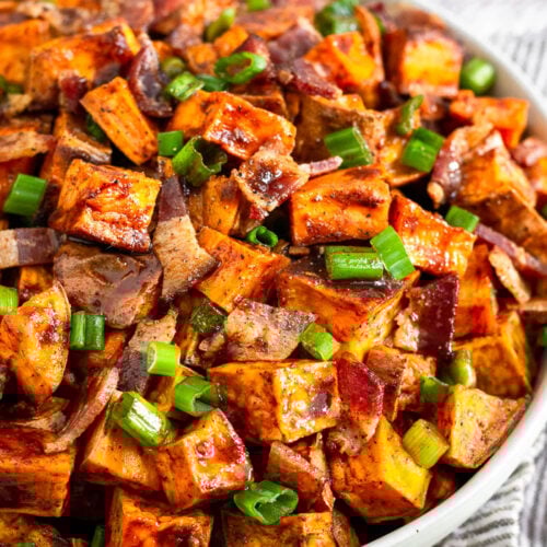 Bowl of glazed sweet potatoes topped with bacon and green onions. Next to is a bowl of green onions and behind it a glass of whiskey.