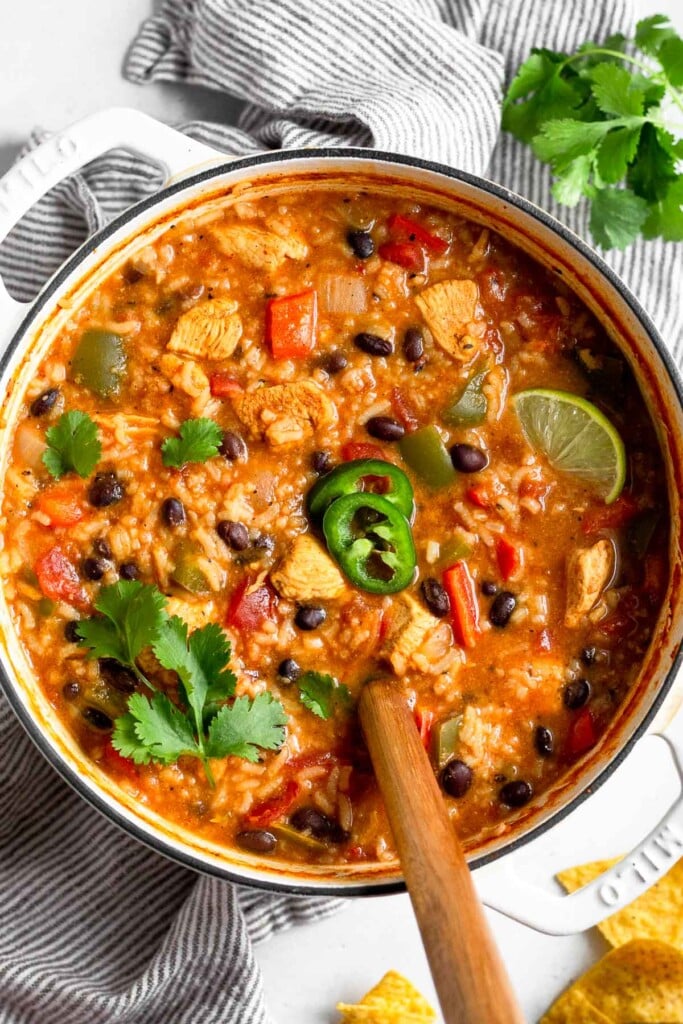 Large pot of chicken fajita soup topped with cilantro, sliced jalapeños, and a lime wedge with a spoon coming out of the pot. The pot is sitting on a striped kitchen towel and around it is a bunch of cilantro and some tortilla chips.
