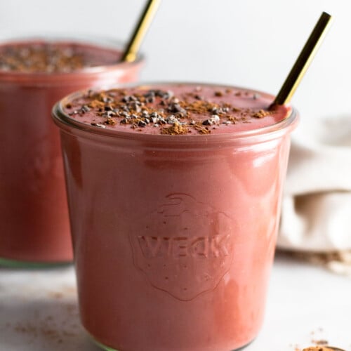 Beet smoothie in a jar topped with cacao nibs and a straw in it. In front of it is a teaspoon filled with cacao powder. Behind it is another jar filled with smoothie.