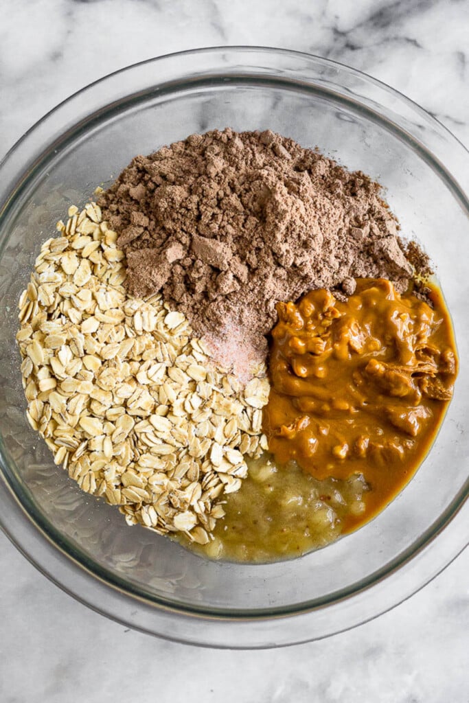 Glass bowl filled with chocolate protein powder, peanut butter, mashed bananas, and rolled oats before it is mixed together.