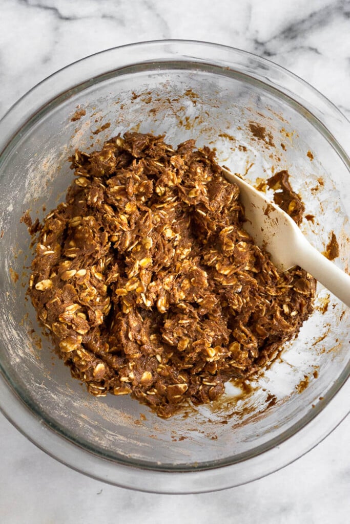 Chocolate dough with oats in it in a large glass bowl. There is also a spatula in the bowl.