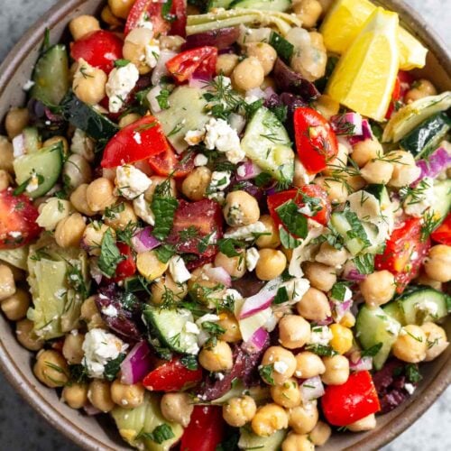 Large bowl filled with greek chickpea salad garnished with fresh herbs and two lemon wedges. Next to it is an empty jar and a bowl full of herbs.