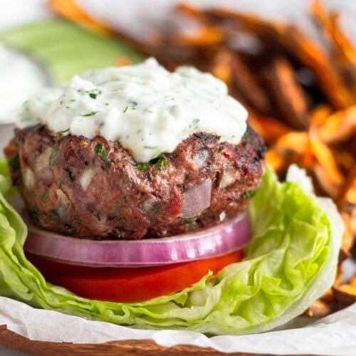 Lamb burger sitting on a lettuce cup with sliced tomato, red onion, and topped with tzatziki sauce. Behind the burger are sweet potatoes fries, sliced cucumbers, and a dish of tzatziki sauce.