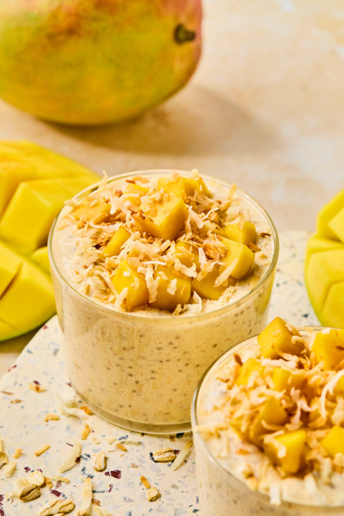 Two jars of blended mango overnight oats topped with diced mango and shredded coconut. Around the jars are some pieces of cut mango attached to the skin and a whole mango.