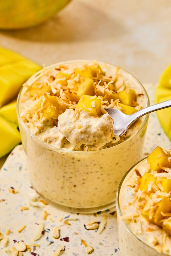 Overnight mango oats in a jar topped with diced mango and shredded coconut. A spoon is being scooped out of the jar with overnight oats on it. Around the jar is another jar of overnight oats and some pieces of cut mango attached to the skin.